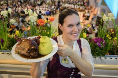 Eine Frau hält einen Teller mit Fleisch und Knödeln, hinter ihr sind Blumen zu sehen.