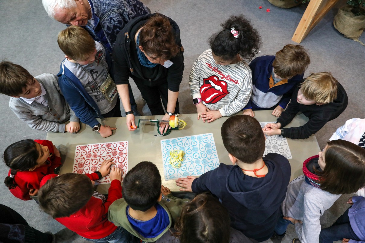 Children stand around a table, watching a device for peeling apples.